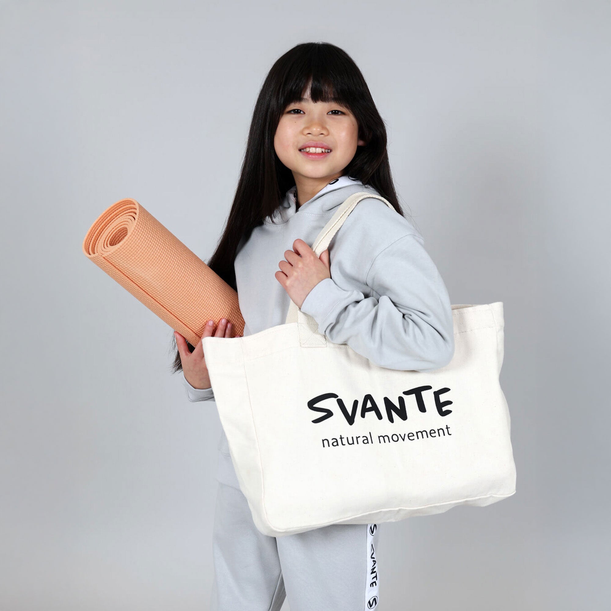 Young Chinese girl with grey organic tracksuit holding a yoga mat and  an organic cotton tote bag with 'SVANTE natural movement' branding on a grey background. 