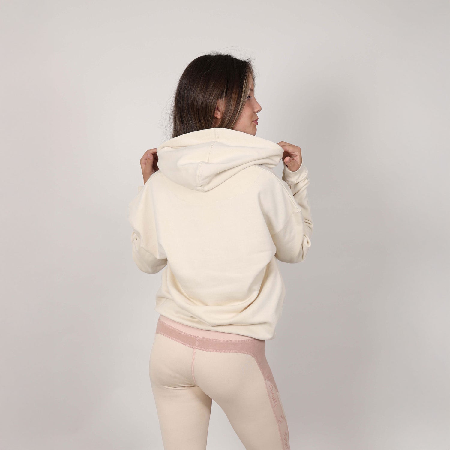 Teenage girl with back to camera showing reverse side of a cream-coloured hoodie and sports leggings on a muted background