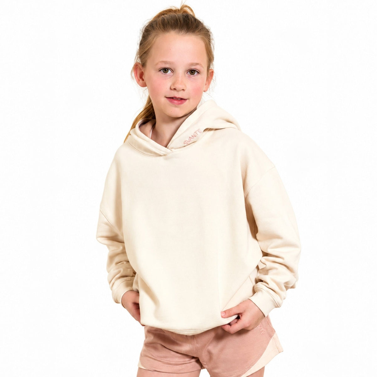 A young girl with blonde hair in a ponytail wearing a Svante organic cotton  cream-coloured oversized hoodie and light pink shorts, standing against a solid white background.