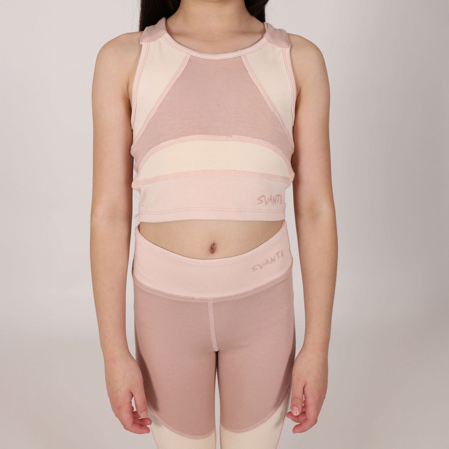 Close up front shot of 8 year old girl wearing a pink and beige athletic outfit with 'Svante' branding on a plain background