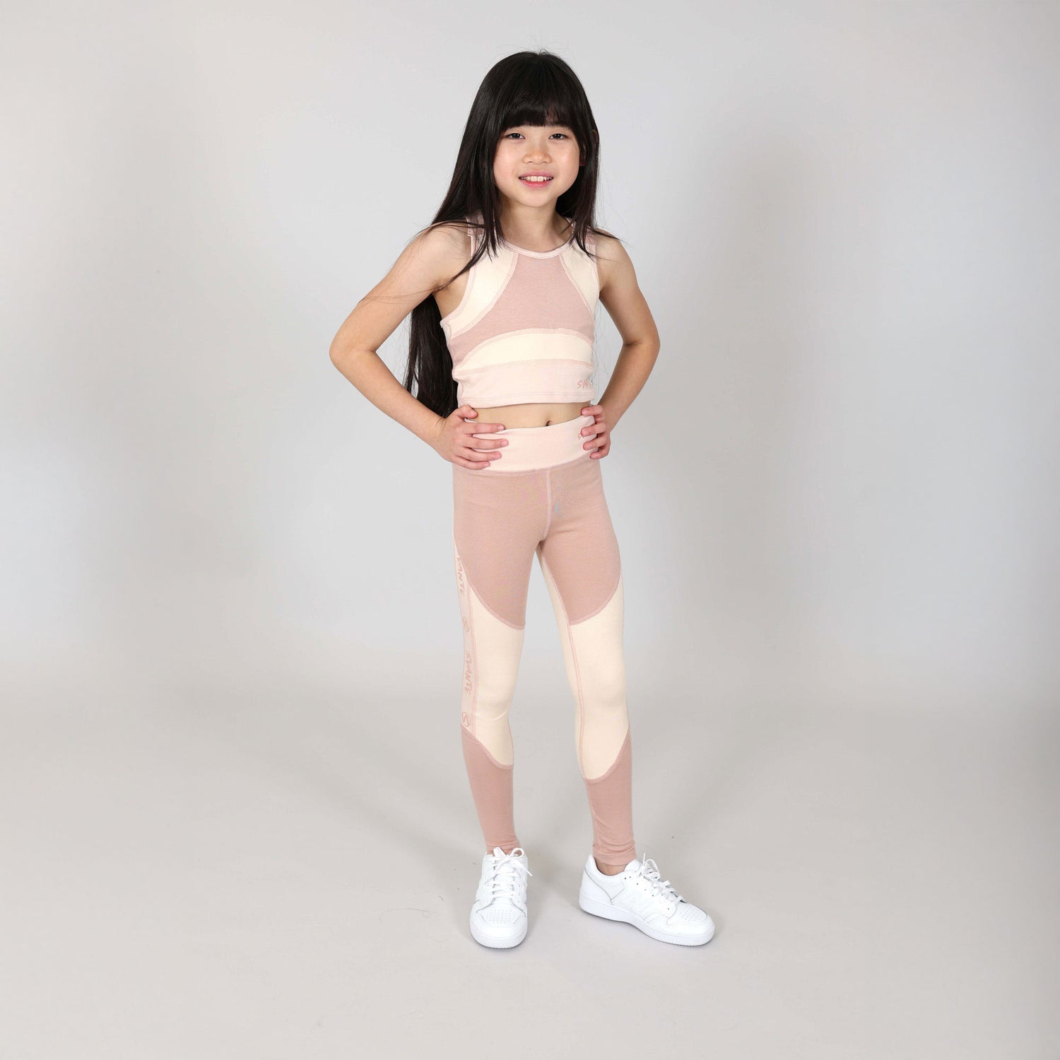 Young girl wearing a matching pink and beige sports crop top and leggings with white sneakers on a plain background
