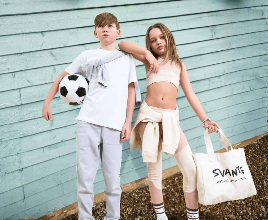 A boy in grey organic cotton joggers holding a football, next to a girl in a plant-based crop top with a hoodie tied at her waist
