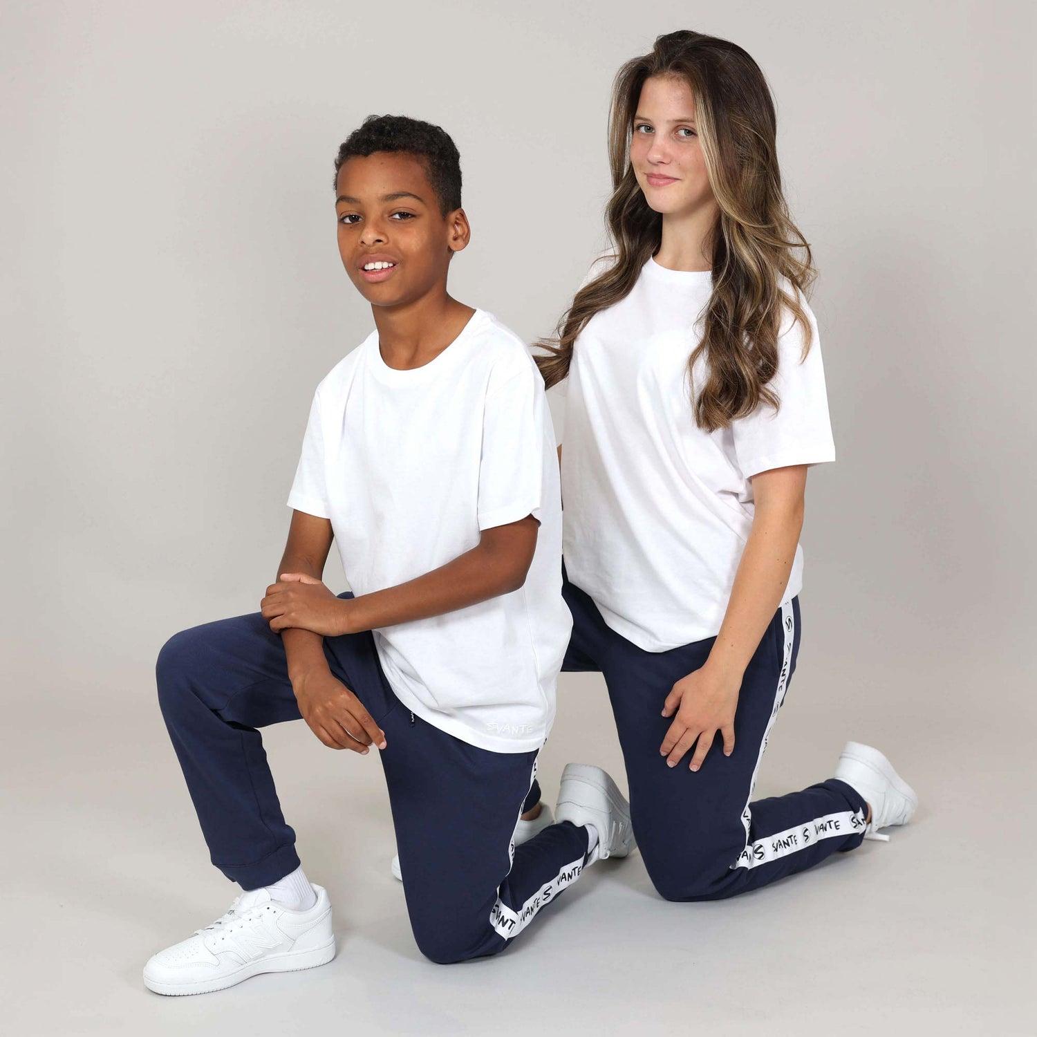 Boy and girl modelling unisex matching outfits of organic white t-shirts and navy tracksuit bottoms with white trainers on a plain background. 