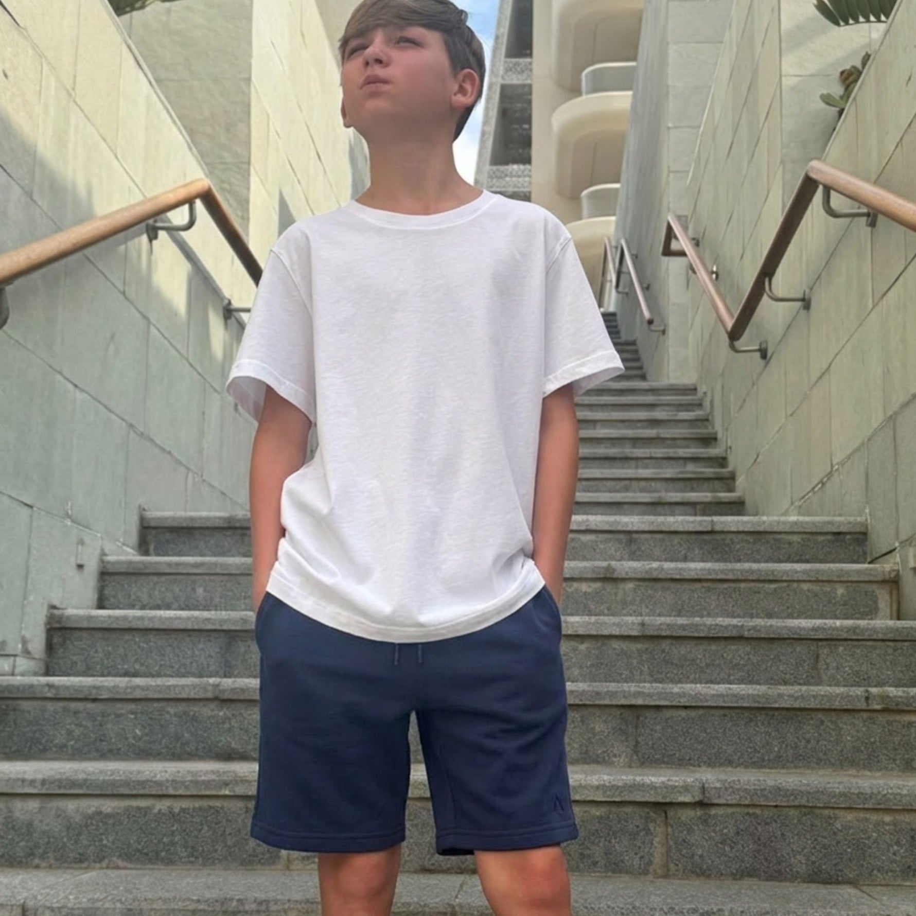 Boy wearing a white organic cotton t-shirt and blue organic cotton jersey shorts standing on stone steps. 